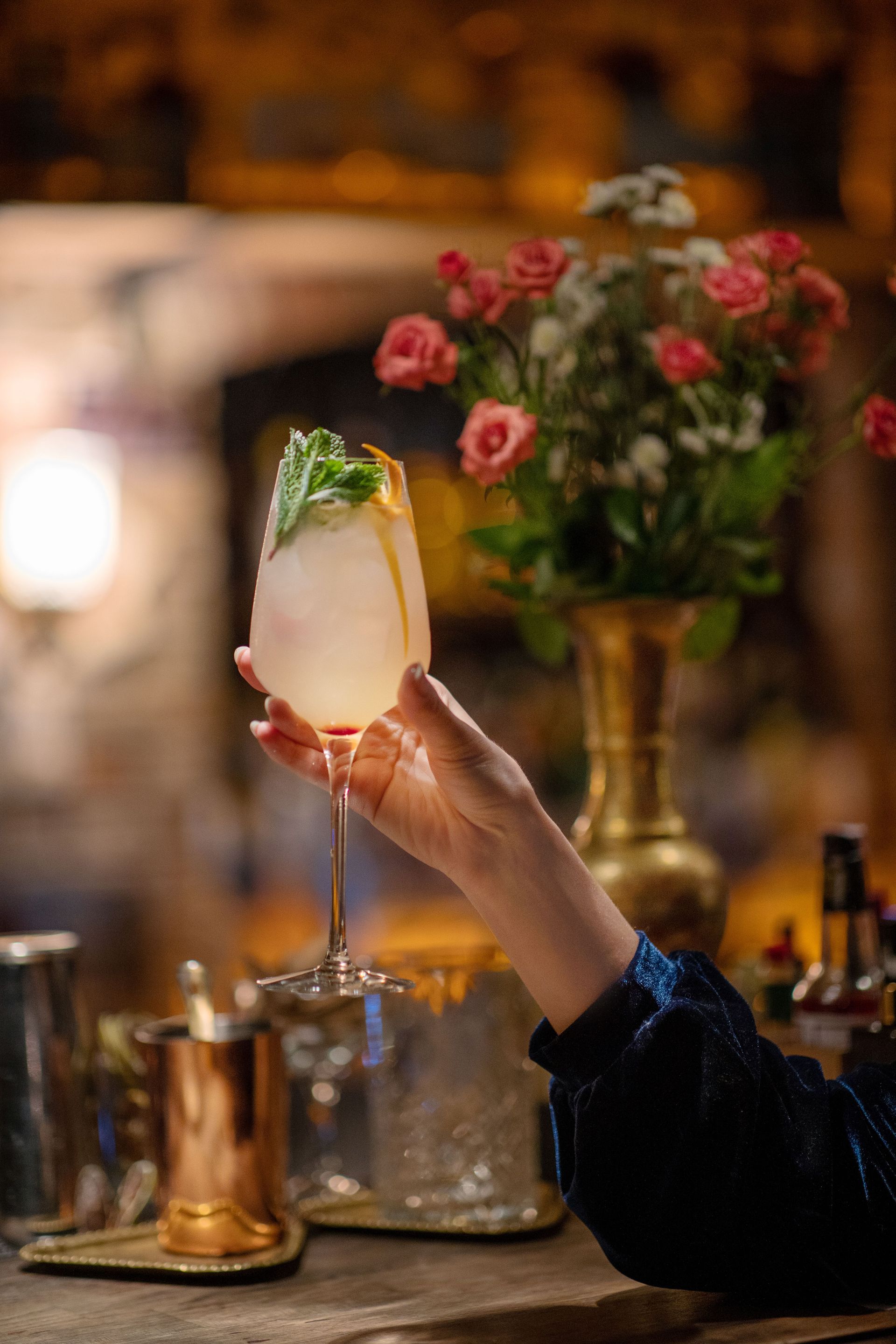 A woman's hand elegantly holds a sophisticated cocktail with mint garnish in a dimly lit bar setting.